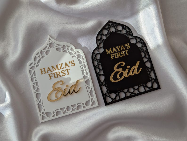 Peut inclure: Deux plaques d&eacute;coratives pour l'A&iuml;d, une blanche et une noire, avec des lettres dor&eacute;es. La plaque blanche indique "Hamza's First Eid", et la plaque noire indique "Maya's First Eid". Les deux plaques ont un design en forme d'arche orn&eacute;.
