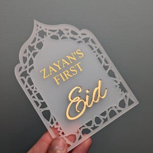 Peut inclure: Plaque d&eacute;corative en acrylique givr&eacute; avec une inscription dor&eacute;e. Le texte "Zayan's First Eid" est &eacute;crit dans une police stylis&eacute;e. La plaque a une bordure d&eacute;corative et est pr&eacute;sent&eacute;e sur un fond gris.