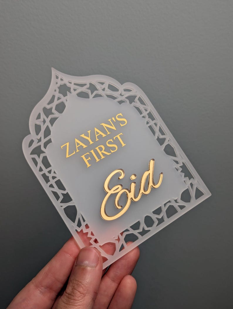 Peut inclure: Plaque d&eacute;corative en acrylique givr&eacute; avec une inscription dor&eacute;e. Le texte "Zayan's First Eid" est &eacute;crit dans une police stylis&eacute;e. La plaque a une bordure d&eacute;corative et est pr&eacute;sent&eacute;e sur un fond gris.