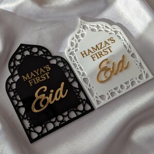 Peut inclure: Deux plaques d&eacute;coratives de l'A&iuml;d, une noire et une blanche, avec des motifs d&eacute;coup&eacute;s complexes. La plaque noire porte l'inscription "MAYA'S FIRST Eid" en lettres dor&eacute;es. La plaque blanche porte l'inscription "HAMZA'S FIRST Eid" en lettres dor&eacute;es. Les plaques sont sur une surface en satin blanc.
