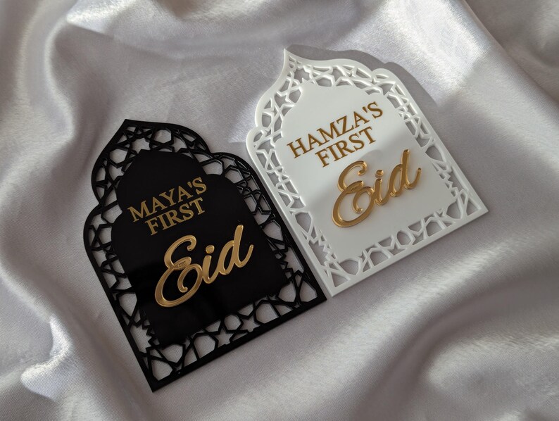 Peut inclure: Deux plaques d&eacute;coratives de l'A&iuml;d, une noire et une blanche, avec des motifs d&eacute;coup&eacute;s complexes. La plaque noire porte l'inscription "MAYA'S FIRST Eid" en lettres dor&eacute;es. La plaque blanche porte l'inscription "HAMZA'S FIRST Eid" en lettres dor&eacute;es. Les plaques sont sur une surface en satin blanc.