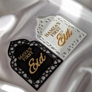Peut inclure: Deux plaques d&eacute;coratives pour l'A&iuml;d, une blanche et une noire, avec des lettres dor&eacute;es. La plaque blanche indique "Hamza's First Eid", et la plaque noire indique "Maya's First Eid". Les deux plaques ont une bordure d&eacute;corative.