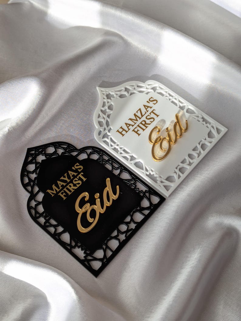 Peut inclure: Deux plaques d&eacute;coratives pour l'A&iuml;d, une blanche et une noire, avec des lettres dor&eacute;es. La plaque blanche indique "Hamza's First Eid", et la plaque noire indique "Maya's First Eid". Les deux plaques ont une bordure d&eacute;corative.