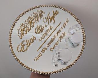 May include: A round, mirrored tray with gold text and a gold beaded border. The text includes names, a date, and a quote. Three clear, faceted candle holders are on the tray. The tray is a wedding or event decoration.