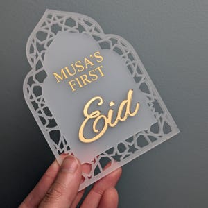 Peut inclure: Plaque d&eacute;corative en acrylique blanc avec l'inscription dor&eacute;e "Musa's First Eid". Le contour est orn&eacute; d'un motif g&eacute;om&eacute;trique ajour&eacute;, et le centre est givr&eacute;. La forme g&eacute;n&eacute;rale &eacute;voque une fen&ecirc;tre cintr&eacute;e, id&eacute;ale pour une c&eacute;l&eacute;bration.