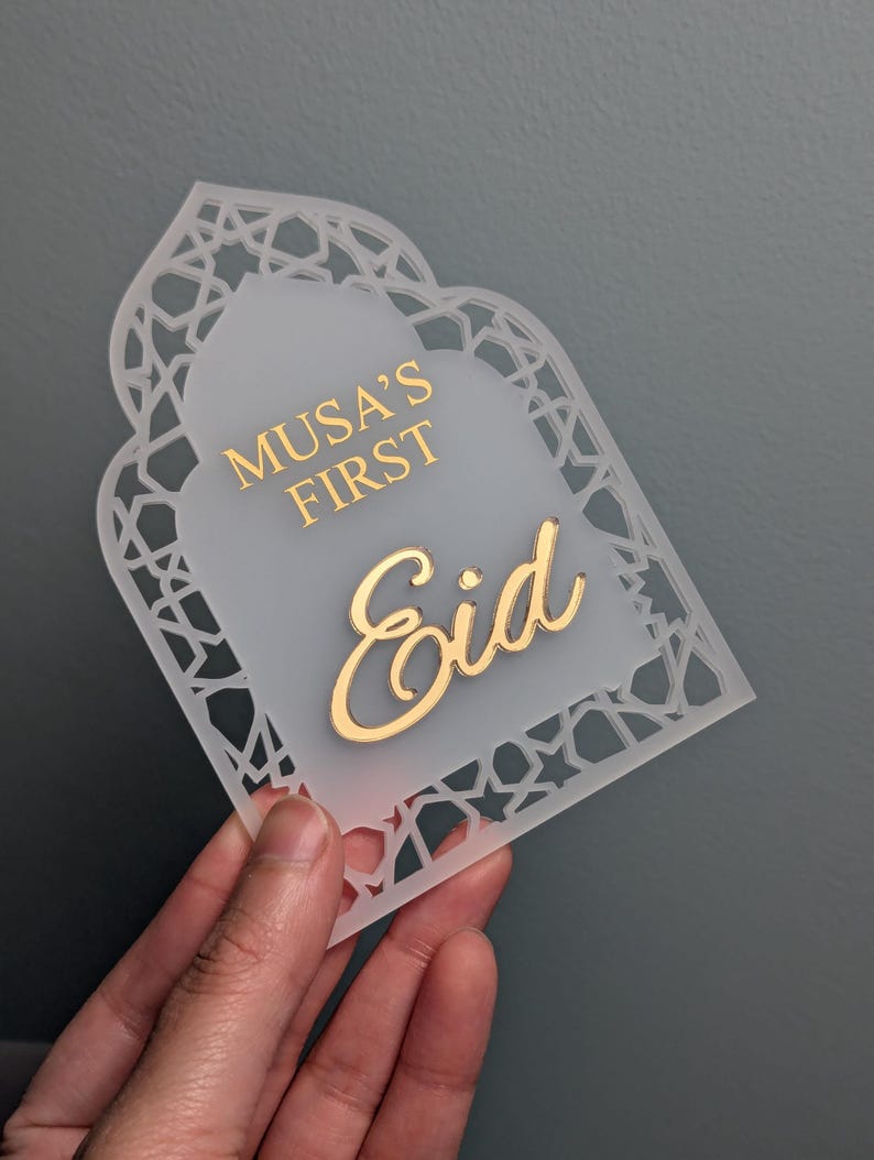 Peut inclure: Plaque d&eacute;corative en acrylique blanc avec l'inscription dor&eacute;e "Musa's First Eid". Le contour est orn&eacute; d'un motif g&eacute;om&eacute;trique ajour&eacute;, et le centre est givr&eacute;. La forme g&eacute;n&eacute;rale &eacute;voque une fen&ecirc;tre cintr&eacute;e, id&eacute;ale pour une c&eacute;l&eacute;bration.