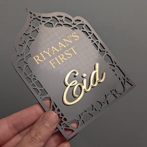 Peut inclure: Plaque d&eacute;corative grise avec une bordure dor&eacute;e et un design d&eacute;coup&eacute; complexe. La plaque porte l'inscription "Riyaan's First Eid" en lettres dor&eacute;es. La plaque est tenue par une main sur un fond gris.