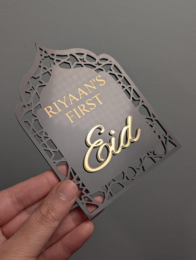Peut inclure: Plaque d&eacute;corative grise avec une bordure dor&eacute;e et un design d&eacute;coup&eacute; complexe. La plaque porte l'inscription "Riyaan's First Eid" en lettres dor&eacute;es. La plaque est tenue par une main sur un fond gris.