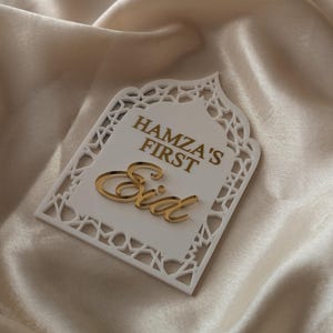 Peut inclure: Plaque d&eacute;corative blanche d&eacute;coup&eacute;e au laser avec l'inscription dor&eacute;e "HAMZA'S FIRST Eid". La plaque a une bordure d&eacute;corative et est pos&eacute;e sur un fond satin&eacute;.