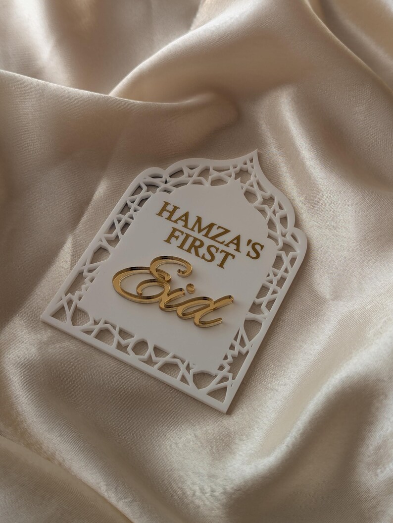 Peut inclure: Plaque d&eacute;corative blanche d&eacute;coup&eacute;e au laser avec l'inscription dor&eacute;e "HAMZA'S FIRST Eid". La plaque a une bordure d&eacute;corative et est pos&eacute;e sur un fond satin&eacute;.
