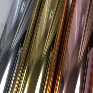 May include: Close-up of three rolls of metallic foil in silver, gold, and rose gold. The foil reflects light, creating a shiny, reflective surface. The rolls are positioned diagonally, showcasing the different colours and textures.