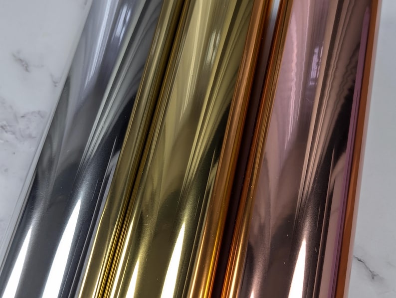 May include: Close-up of three rolls of metallic foil in silver, gold, and rose gold. The foil reflects light, creating a shiny, reflective surface. The rolls are positioned diagonally, showcasing the different colours and textures.