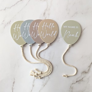 May include: Five pastel-coloured balloon-shaped decorations with white text. Four balloons say "Hello World" and one says "My Name Is Noah". Each balloon has a white rope attached. The balloons are in shades of green, blue, and tan.