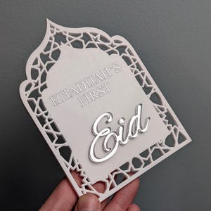 Peut inclure: Plaque d&eacute;corative blanche d&eacute;coup&eacute;e au laser avec une bordure g&eacute;om&eacute;trique complexe. Le texte "KHADIJAH'S FIRST" et le mot "Eid" en argent sont pr&eacute;sents. La plaque est tenue &agrave; la main sur un fond gris.