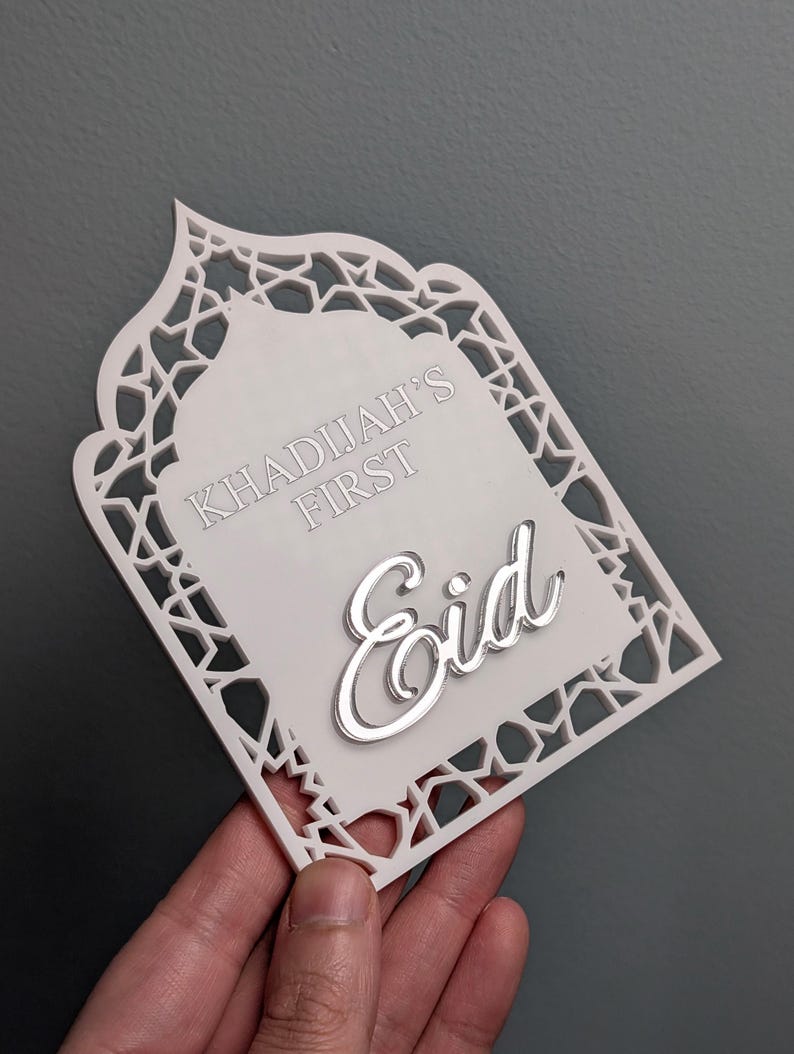 Peut inclure: Plaque d&eacute;corative blanche d&eacute;coup&eacute;e au laser avec une bordure g&eacute;om&eacute;trique complexe. Le texte "KHADIJAH'S FIRST" et le mot "Eid" en argent sont pr&eacute;sents. La plaque est tenue &agrave; la main sur un fond gris.