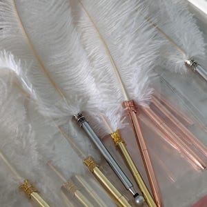 May include: A selection of elegant pens in gold, silver, and rose gold, each with a decorative crown-shaped top. The pens are displayed alongside large, fluffy white feathers. The pens are presented on a clear surface.
