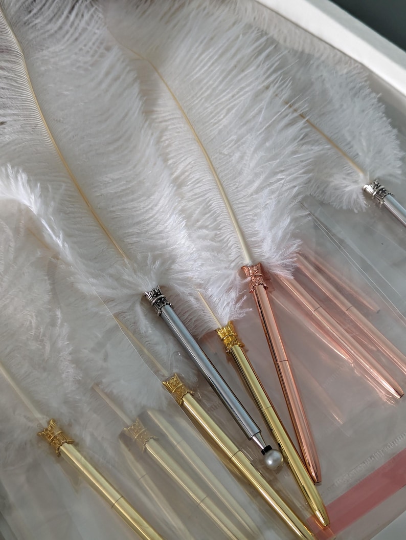 May include: A selection of elegant pens in gold, silver, and rose gold, each with a decorative crown-shaped top. The pens are displayed alongside large, fluffy white feathers. The pens are presented on a clear surface.