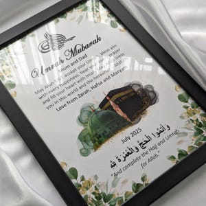 May include: A black framed print with the words "Umrah Mubarak" and Arabic script. The print includes illustrations of religious sites, floral designs, and the text "July 2025".