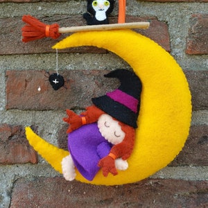 May include: A felt witch doll sleeping on a yellow crescent moon with a black cat on a broom above. The witch is wearing a purple dress and a black hat with a purple band.