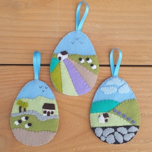 May include: Three felt ornaments shaped like eggs, each depicting a different countryside scene. The first ornament features a white house with a blue roof, green fields, and a purple sky. The second ornament shows a white house with a brown roof, green hills, and a blue sky. The third ornament has a white house with a blue roof, green hills, and a blue sky with clouds.