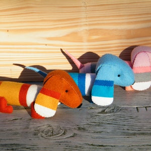May include: Four felt dachshund dog toys in different colors and patterns. The dogs are orange and white, blue and white, pink and gray, and blue and white. The dogs are lying on a wooden surface.