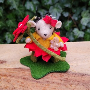 Puede incluir: Un erizo de fieltro que lleva un disfraz de poinsettia roja y sostiene una persona de jengibre. El erizo está de pie sobre una estrella de fieltro verde.