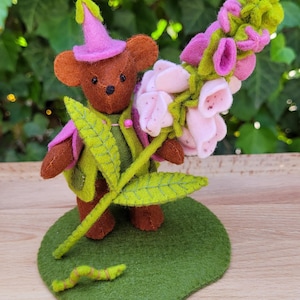 May include: A handmade felt teddy bear wearing a pink hat and green outfit, holding a bouquet of pink and green flowers. The bear stands on a green felt base with a green felt leaf and worm. The background is blurred green foliage.