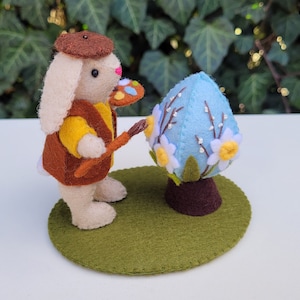 May include: A felt bunny artist wearing a brown beret, yellow shirt, and brown vest, painting a light blue Easter egg decorated with white and yellow flowers. The bunny stands on a green felt base, creating a whimsical Easter scene.