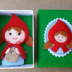 May include: A felt craft of Little Red Riding Hood holding a basket. The craft is displayed in a white box with a green felt background. The box is closed with a green felt lid with a felt craft of Little Red Riding Hood on it.