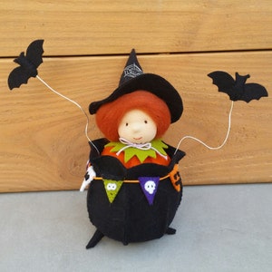 May include: A felt witch doll with red hair wearing a black witch hat with a spider web design. The doll is sitting inside a black cauldron with a pumpkin face and Halloween-themed flags.