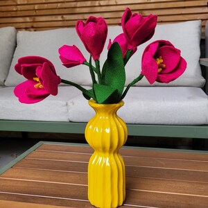 Peut inclure: Un vase en céramique jaune vif contient un bouquet de tulipes en feutre. Les tulipes sont d'un rose profond avec des tiges et des feuilles vertes. Le vase a un design côtelé et repose sur une table en bois.
