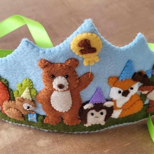 May include: A blue felt crown with a green ribbon, featuring a bear, fox, deer, and squirrel wearing party hats. The crown has a yellow balloon with the number "1" on it.