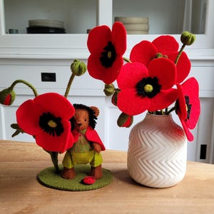 Peut inclure: Une composition décorative avec des coquelicots en feutre. Un vase blanc contient un bouquet de coquelicots rouges à centre noir. Un ours en peluche en feutre avec une cape rouge et une tenue verte se tient près d'un grand coquelicot. Une coccinelle rouge est sur la base verte.