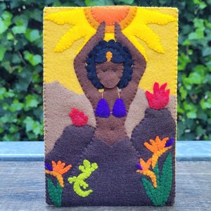 May include: A felt art piece depicting a woman with arms raised towards a yellow sun. The woman wears a purple bikini. Brown mountains and red and orange flowers are at the bottom. A green lizard is also present.