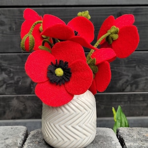 Peut inclure: Un bouquet de coquelicots rouges en feutre, avec des centres noirs et jaunes, présenté dans un petit vase en céramique blanche à motif chevron. Les tiges et les boutons sont verts.