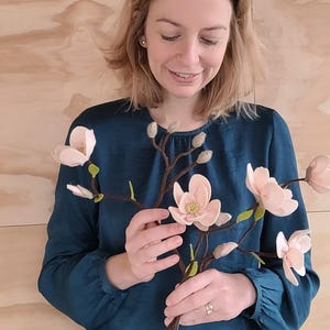 Op de afbeelding: Een persoon houdt een tak met vilten magnoliabloemen vast. De bloemen zijn zachtroze met groene accenten en de stelen zijn bruin. De persoon draagt een tealkleurige blouse en een gestreepte broek. De achtergrond is een licht houten paneel.