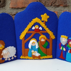 May include: A felt Christmas nativity scene with a blue background. The scene includes Mary, Joseph, baby Jesus, an angel, a shepherd, a sheep, and the three wise men.