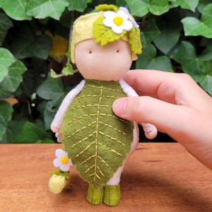 May include: A handmade doll with a leaf-shaped green felt body and a yellow hat adorned with a white daisy. The doll has a beige fabric face and white arms and legs. A small flower bouquet is held in its hand.