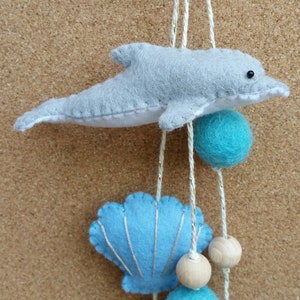 May include: A gray felt dolphin with black eyes hangs from a string with blue and wooden beads and a blue felt seashell.