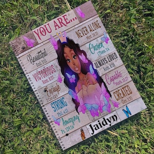May include: A spiral notebook with a wood-grain background and a colorful illustration of a woman with butterflies. The notebook has inspirational quotes and scripture verses, including "You Are... Beautiful, Victorious, Enough, Strong, Amazing, Never Alone, Chosen, Always Loved, Capable, Created". The notebook is personalized with the name "Jaidyn".