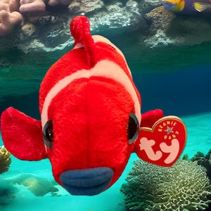 May include: A red and white plush clownfish toy with a blue mouth and large black eyes. The toy has a Ty Beanie Baby tag attached to it. The toy is in front of a blue background with coral and other sea life.