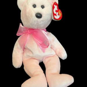 May include: Pink plush bear with a pink ribbon around its neck. The bear has a black nose and black eyes. The bear is sitting on a black background. The bear has a red tag that says "Ty Beanie Babies".