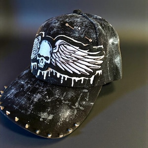 Puede incluir: Gorra de béisbol negra con un acabado desgastado, adornada con un diseño de calavera y alas en blanco y azul claro. La visera presenta tachuelas metálicas. La gorra tiene una estética única y atrevida.