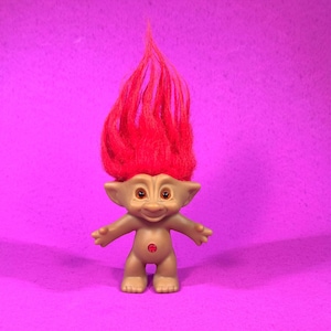 May include: A vintage troll doll with a tan body, small arms, and a red gemstone on its belly. The doll has a smiling face with brown eyes and a large tuft of bright red hair. The background is a solid magenta color.