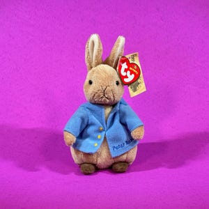 May include: A plush Peter Rabbit toy, wearing a blue jacket with gold buttons, and the name "Peter Rabbit" embroidered on the front. The toy has a Ty tag attached. The background is a solid purple.
