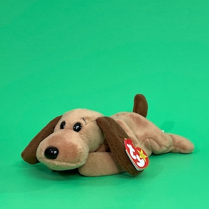 May include: A plush dog toy in shades of brown and tan, with floppy ears and a small tail. The dog has black eyes and a black nose. A red tag with white text is attached to the toy. The background is a solid green.