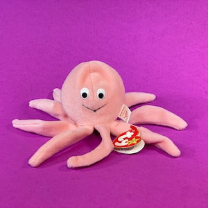 May include: A plush pink octopus toy with a smiling face and eight tentacles. The toy has black eyes and a tag attached. The background is a solid purple color.