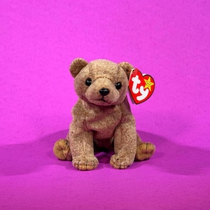 May include: A brown teddy bear plush toy with a red heart-shaped tag. The bear is sitting upright with its paws forward, and has black eyes and a black nose. The background is a solid magenta color.