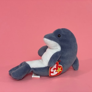 May include: A gray and white plush dolphin toy with a red Beanie Baby tag that says "Ty Inc. Beanie Babies" and has a star on it.