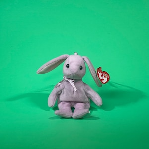 May include: A lavender plush bunny with a white bow and a red "Ty" tag. The bunny is sitting on a green background.